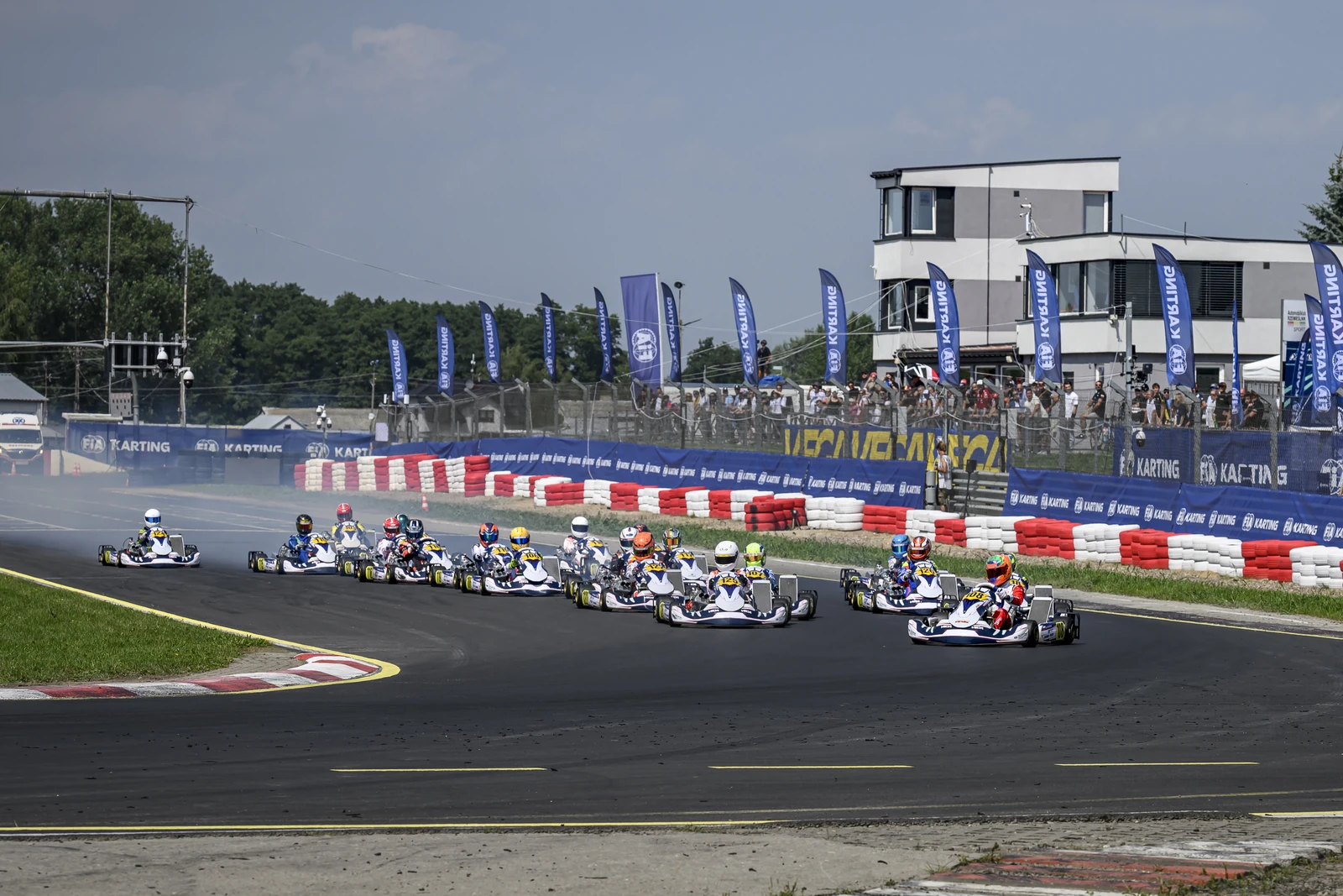 Start wyścigu: peleton identycznych gokartów Kosmic wchodzi w pierwszy zakręt Autodromu Słomczyn, w tle banery FIA Karting